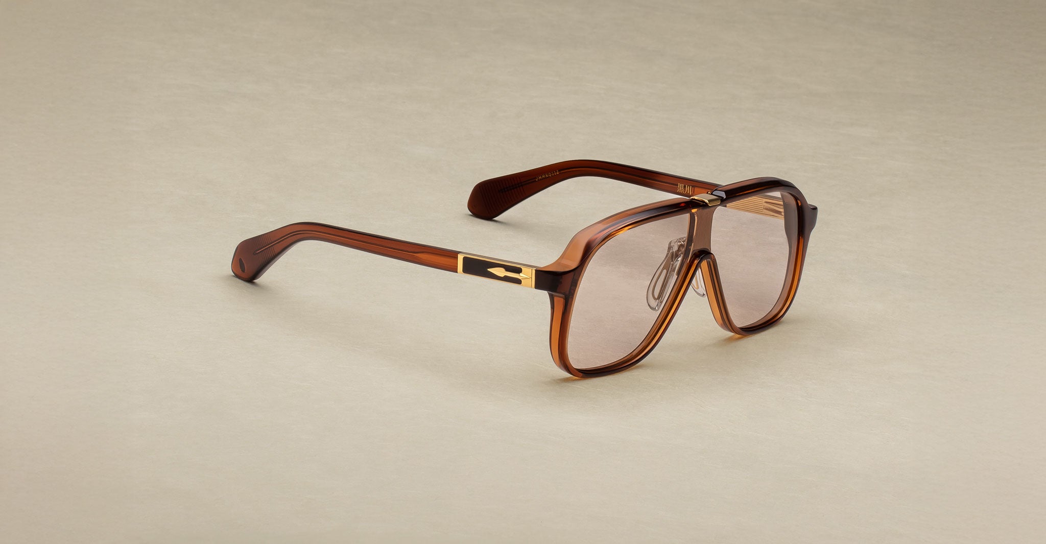 Jacques Marie Mage Kurokawa: Brown aviator-style eyeglasses with a thick frame, gold temple accents, and clear lenses, shown on a beige background.