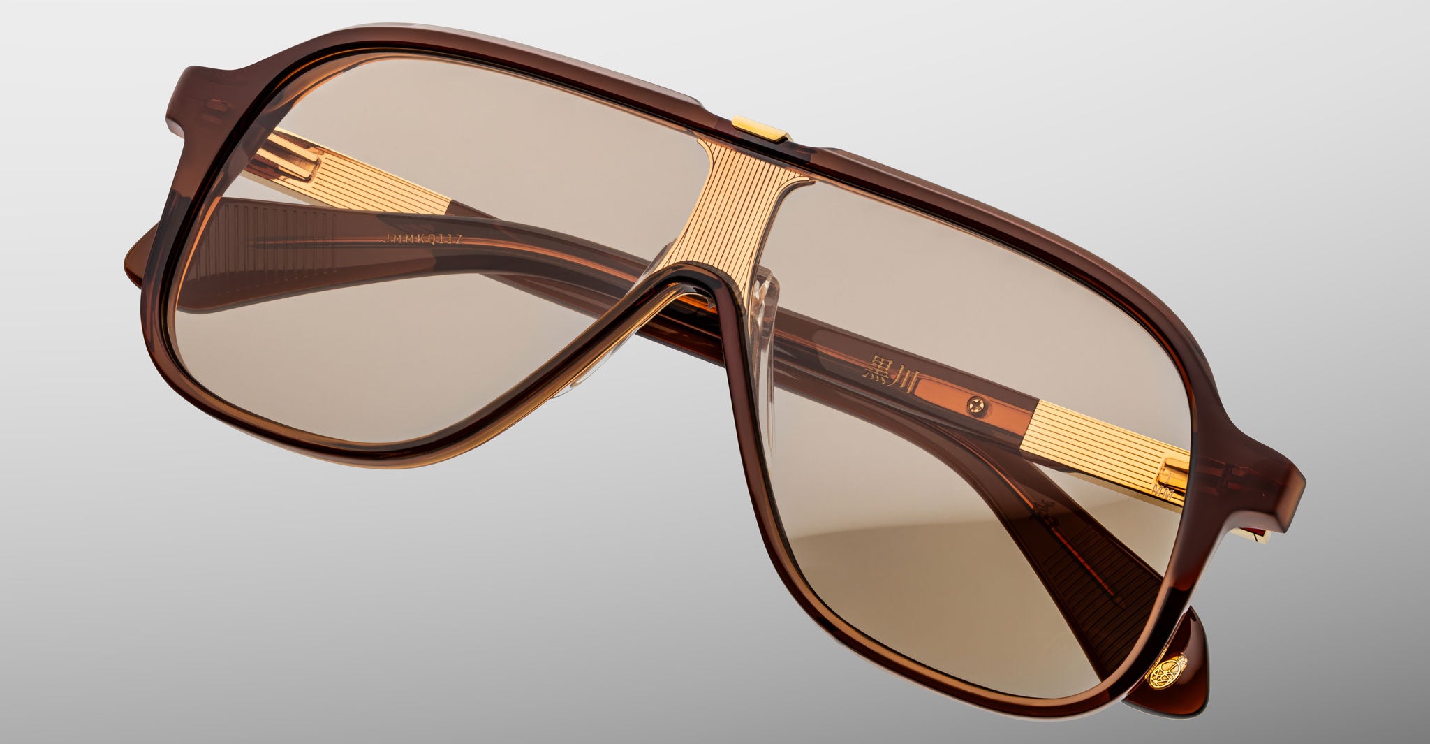 Jacques Marie Mage Kurokawa features brown oversized sunglasses with gold-accented arms and lightly tinted lenses. Showcased on a neutral gray background, these retro, aviator-inspired frames highlight elegant gold detailing.