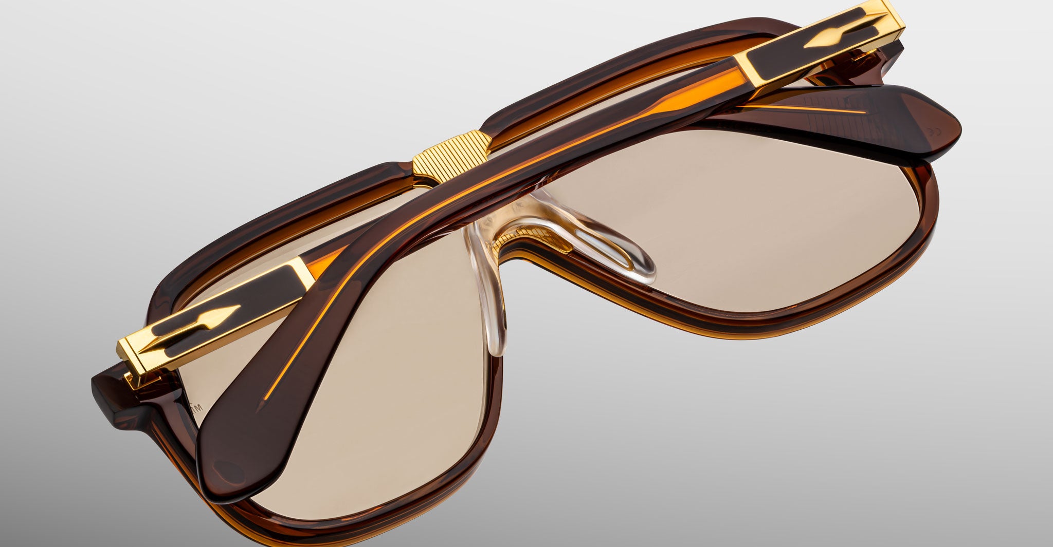 The Jacques Marie Mage Kurokawa sunglasses feature thick brown and gold frames with slightly tinted lenses, displayed from an angled top view on a plain gray background.