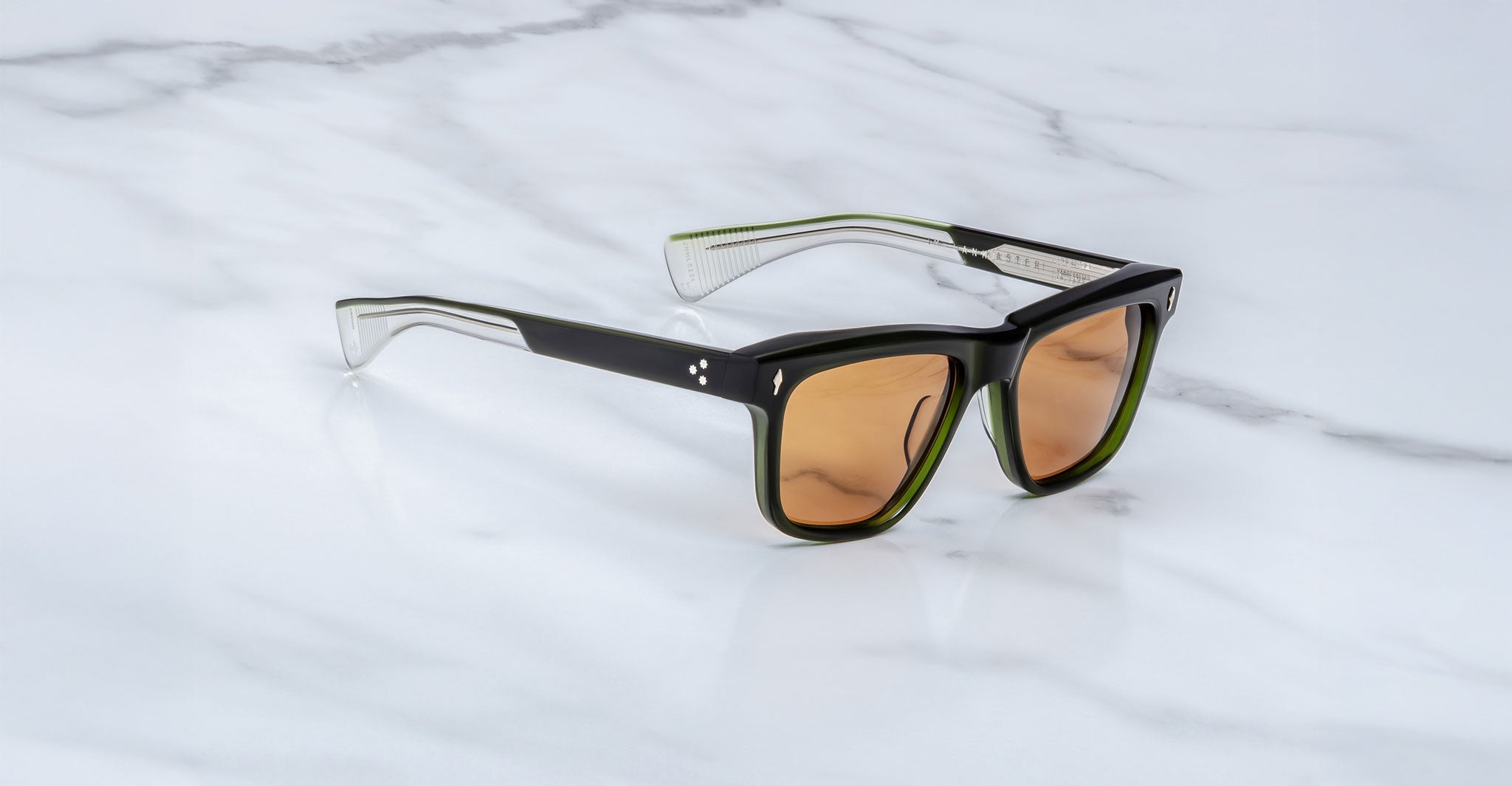 The Jacques Marie Mage Lankaster sunglasses, featuring thick black and transparent frames with brown-tinted lenses, rest on a white marble surface.