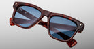 The Jacques Marie Mage Lankaster is a pair of square-framed tortoiseshell sunglasses with blue-tinted lenses, displayed against a plain gray background. The arms feature inscribed branding text.