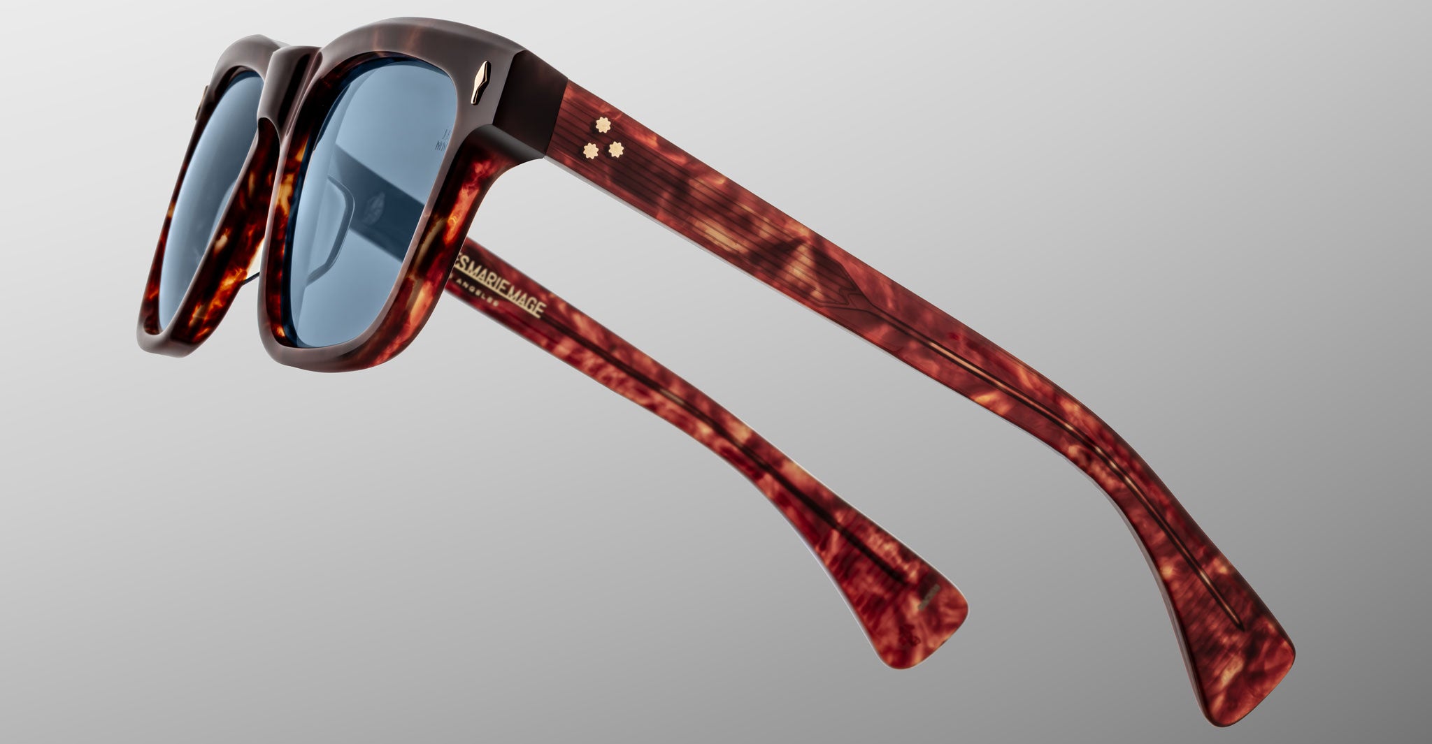 The Jacques Marie Mage Lankaster sunglasses feature a tortoiseshell pattern, blue lenses, thick frames, and gold accents on the arms, shown from a side angle against a plain light gray background.