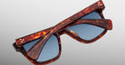 A pair of Jacques Marie Mage Lankaster tortoiseshell sunglasses with blue lenses, folded flat with one temple arm resting atop the other, set against a light gray background.