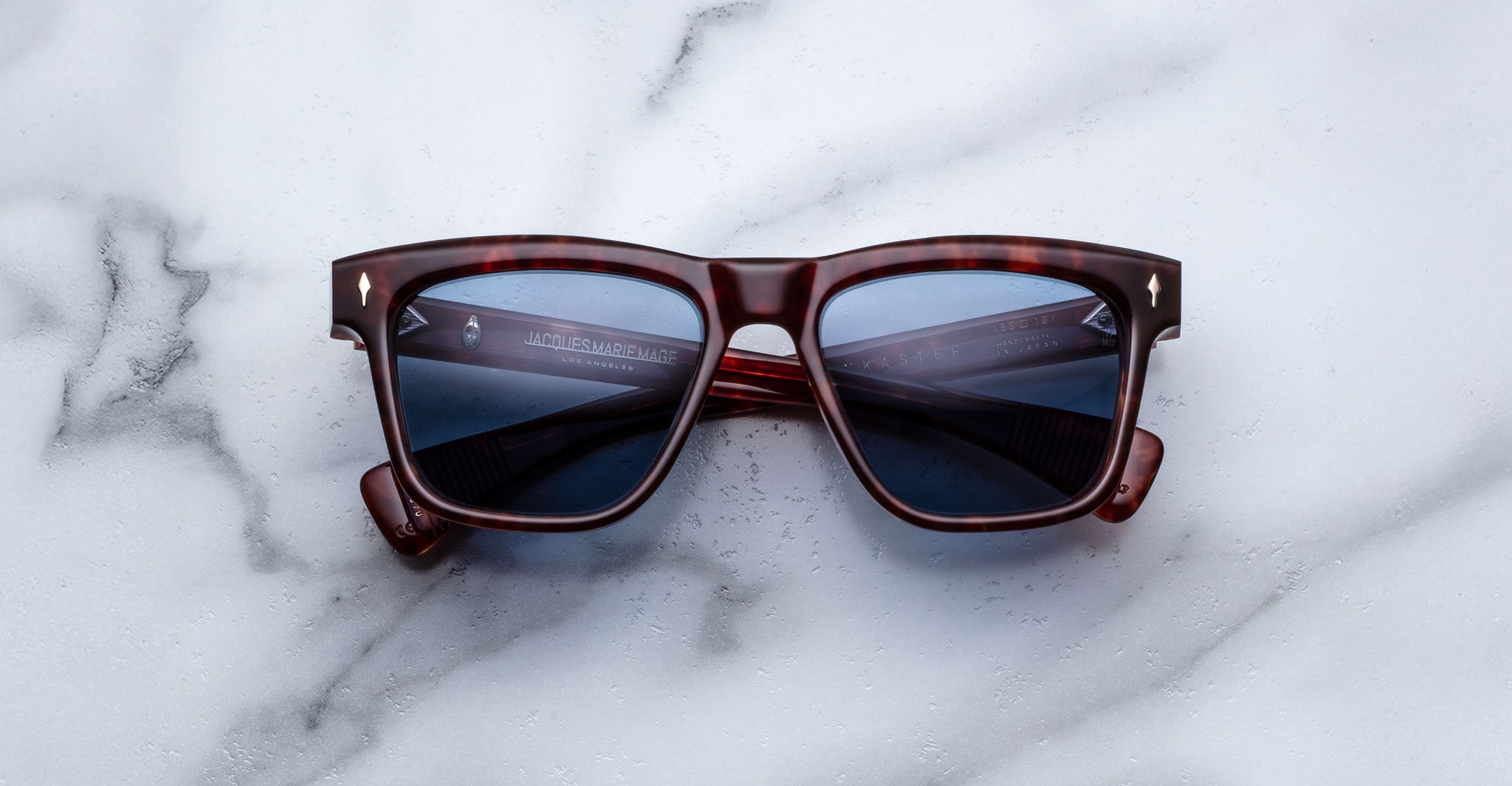 A pair of Jacques Marie Mage Lankaster rectangular tortoiseshell sunglasses with dark lenses rests on a white marble surface.