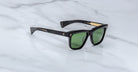 A pair of Jacques Marie Mage Lankaster black sunglasses with green lenses and gold temple accents, displayed on a white marble surface.