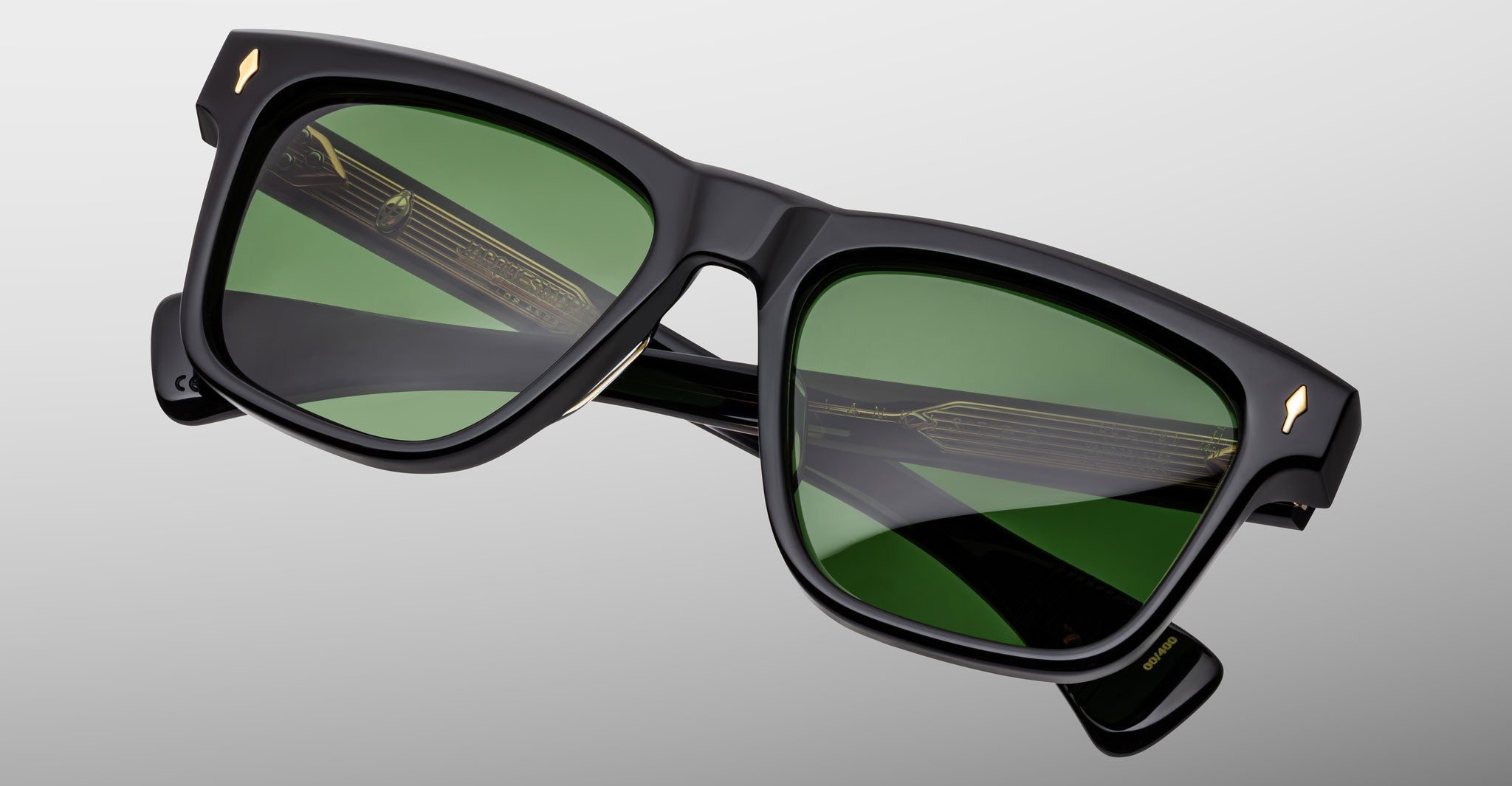 The Jacques Marie Mage Lankaster sunglasses feature a black rectangular frame with green-tinted lenses, gold accents at the front corners, and visible gold detailing on the inner arms, displayed on a light gray background.