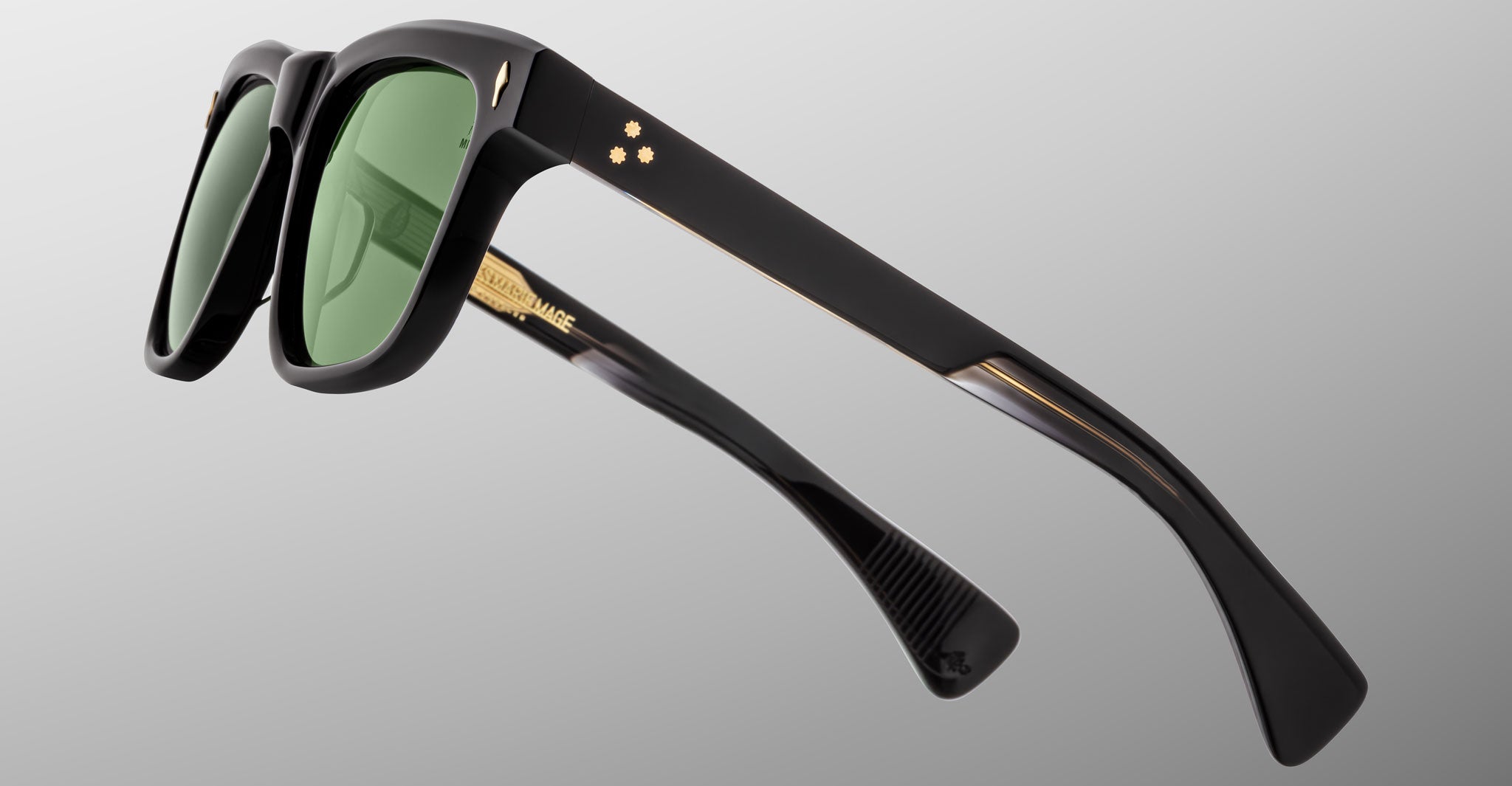 The Jacques Marie Mage Lankaster sunglasses feature black frames with green lenses, gold star-shaped accents, and thick angular arms, displayed against a plain light gray background.