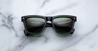 A pair of Jacques Marie Mage Lankaster black rectangular sunglasses with dark green lenses rests on a white marble surface.