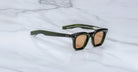 A pair of Jacques Marie Mage Leclair sunglasses with rectangular, dark green frames, brown-tinted lenses, and silver accents by the hinges rests on a white marble surface.