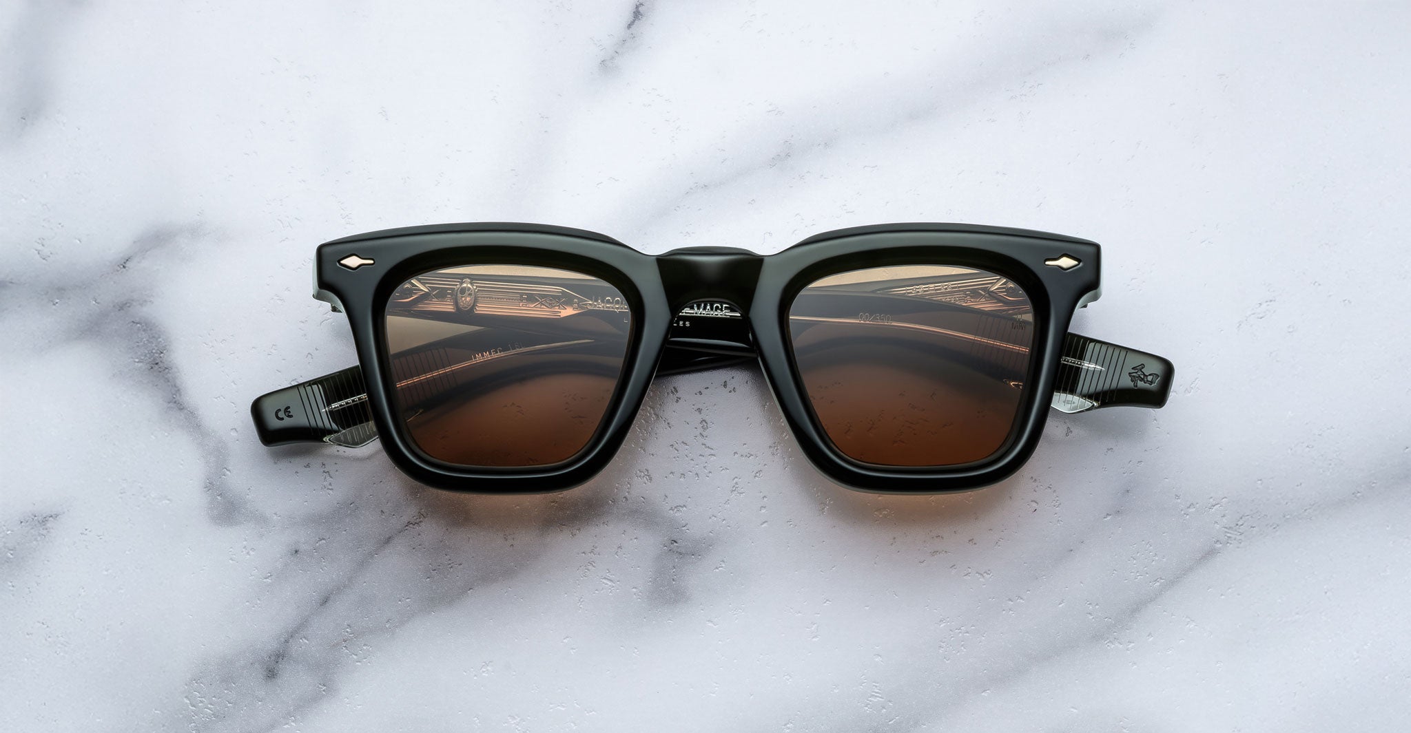 A pair of Jacques Marie Mage Leclair sunglasses with black frames and brown tinted lenses is placed on a white marble surface, viewed from above.