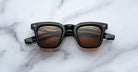 A pair of Jacques Marie Mage Leclair sunglasses with black frames and brown tinted lenses is placed on a white marble surface, viewed from above.
