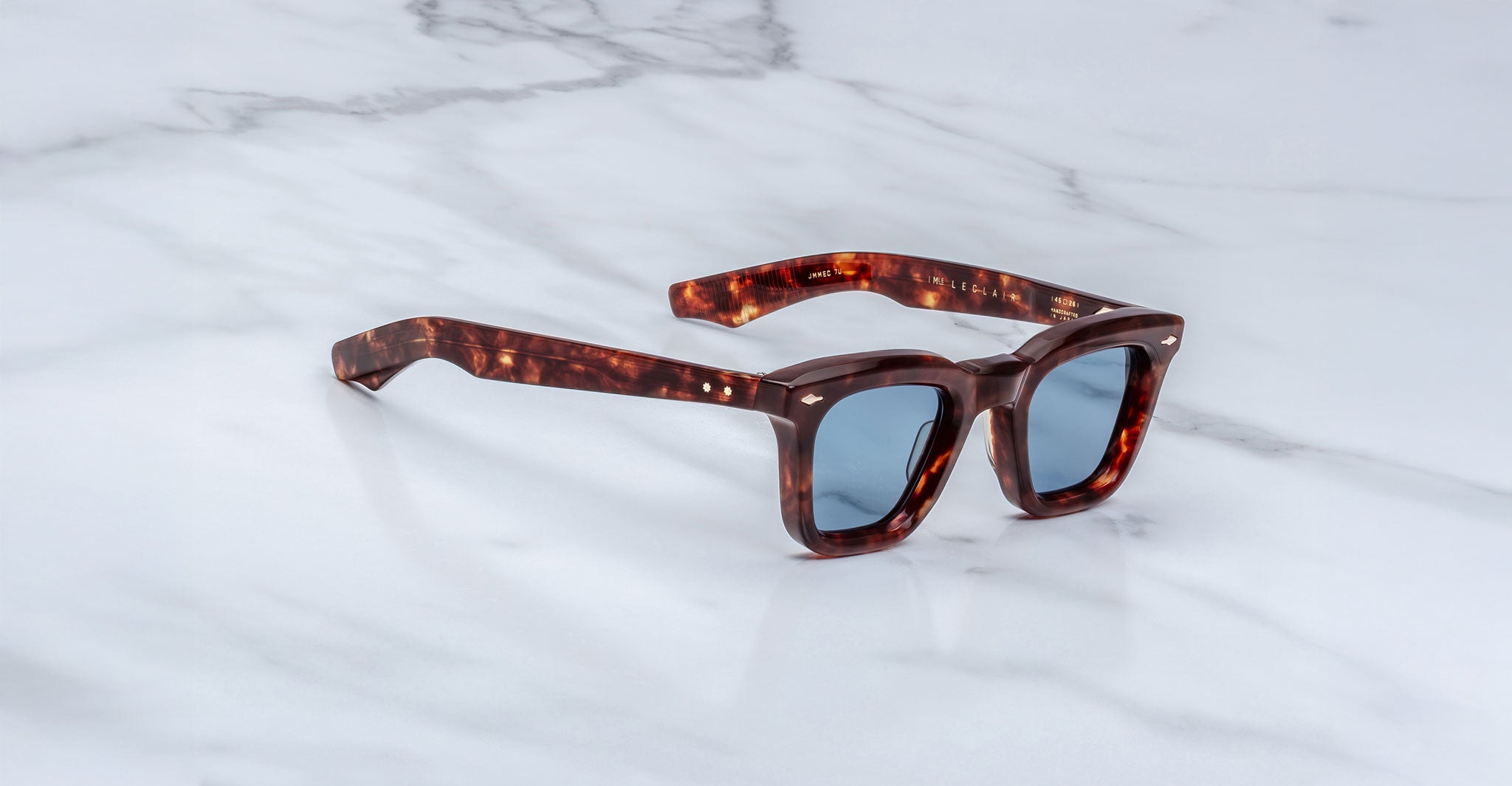 The Jacques Marie Mage Leclair sunglasses, featuring square tortoiseshell frames and blue-tinted lenses, rest on a white marble surface.