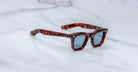 The Jacques Marie Mage Leclair sunglasses, featuring square tortoiseshell frames and blue-tinted lenses, rest on a white marble surface.
