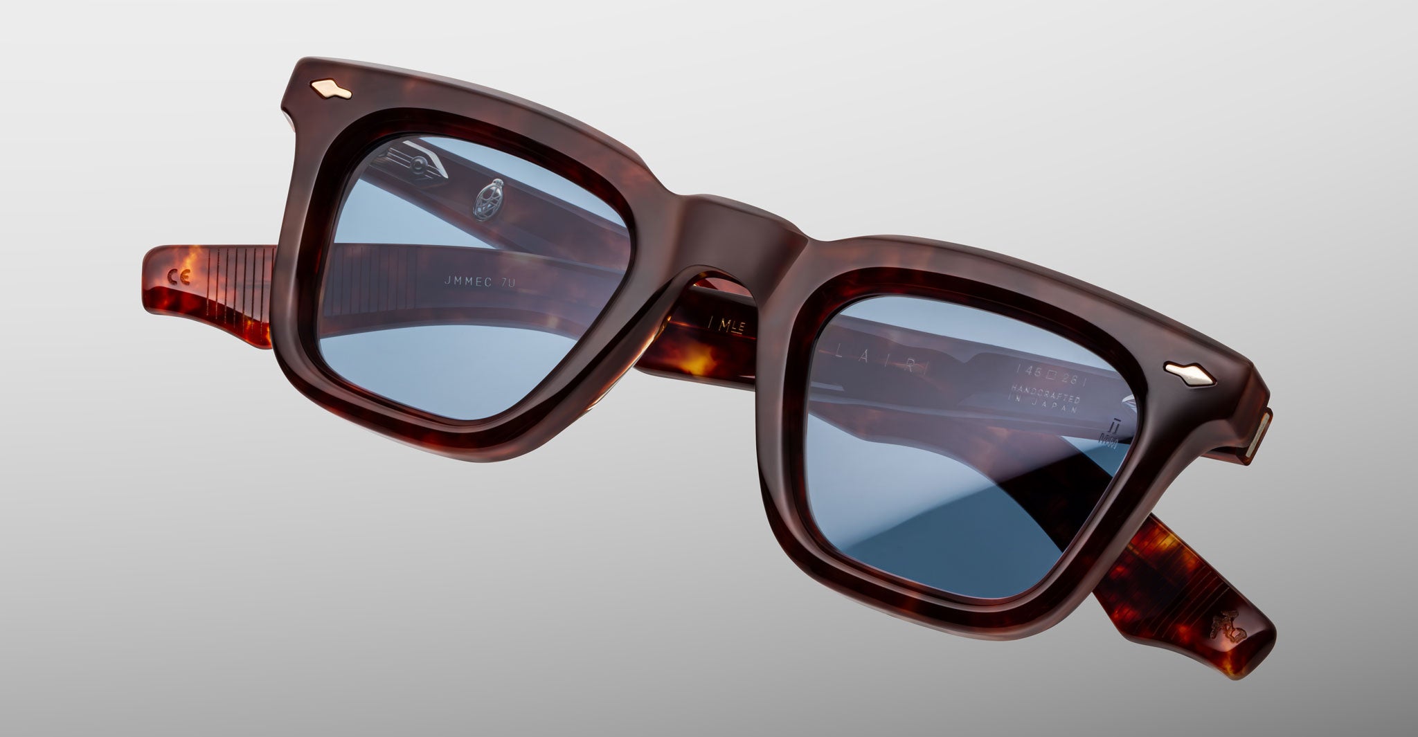 The Jacques Marie Mage Leclair sunglasses feature square frames in dark brown tortoiseshell, blue-tinted reflective lenses, and silver accents on the upper corners, shown against a light gray background.