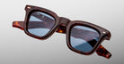 The Jacques Marie Mage Leclair sunglasses feature square frames in dark brown tortoiseshell, blue-tinted reflective lenses, and silver accents on the upper corners, shown against a light gray background.