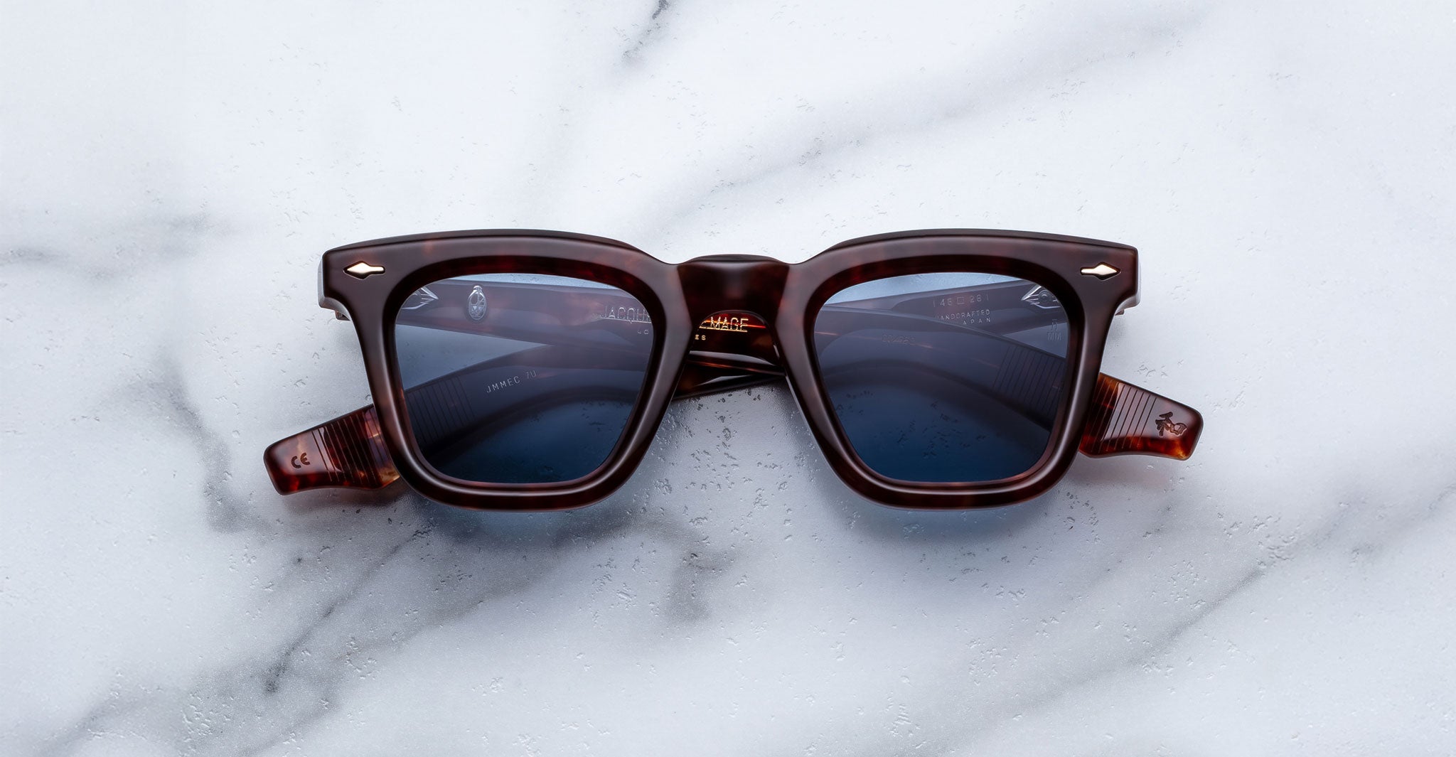 The Jacques Marie Mage Leclair sunglasses, featuring dark brown square frames and blue-tinted lenses, are displayed on a white marble surface.