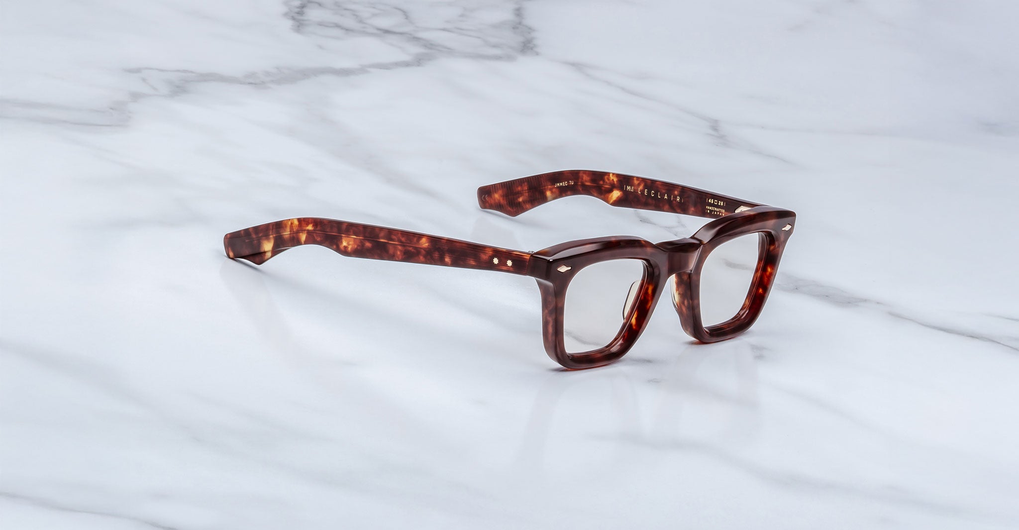 A pair of Jacques Marie Mage Leclair rectangular tortoiseshell eyeglasses with thick frames rests on a white marble surface.