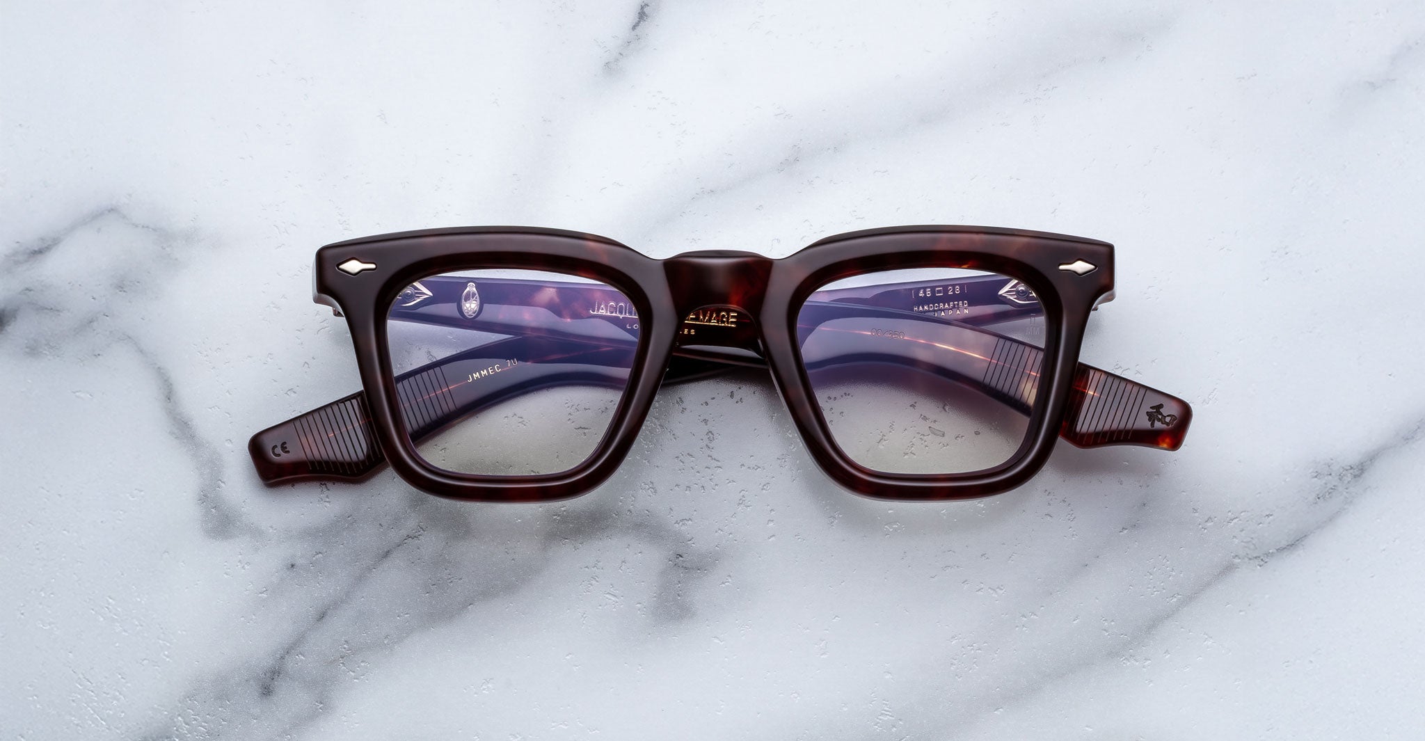 A pair of Jacques Marie Mage Leclair rectangular, thick-framed eyeglasses with a dark tortoiseshell pattern rests on a white marble surface.