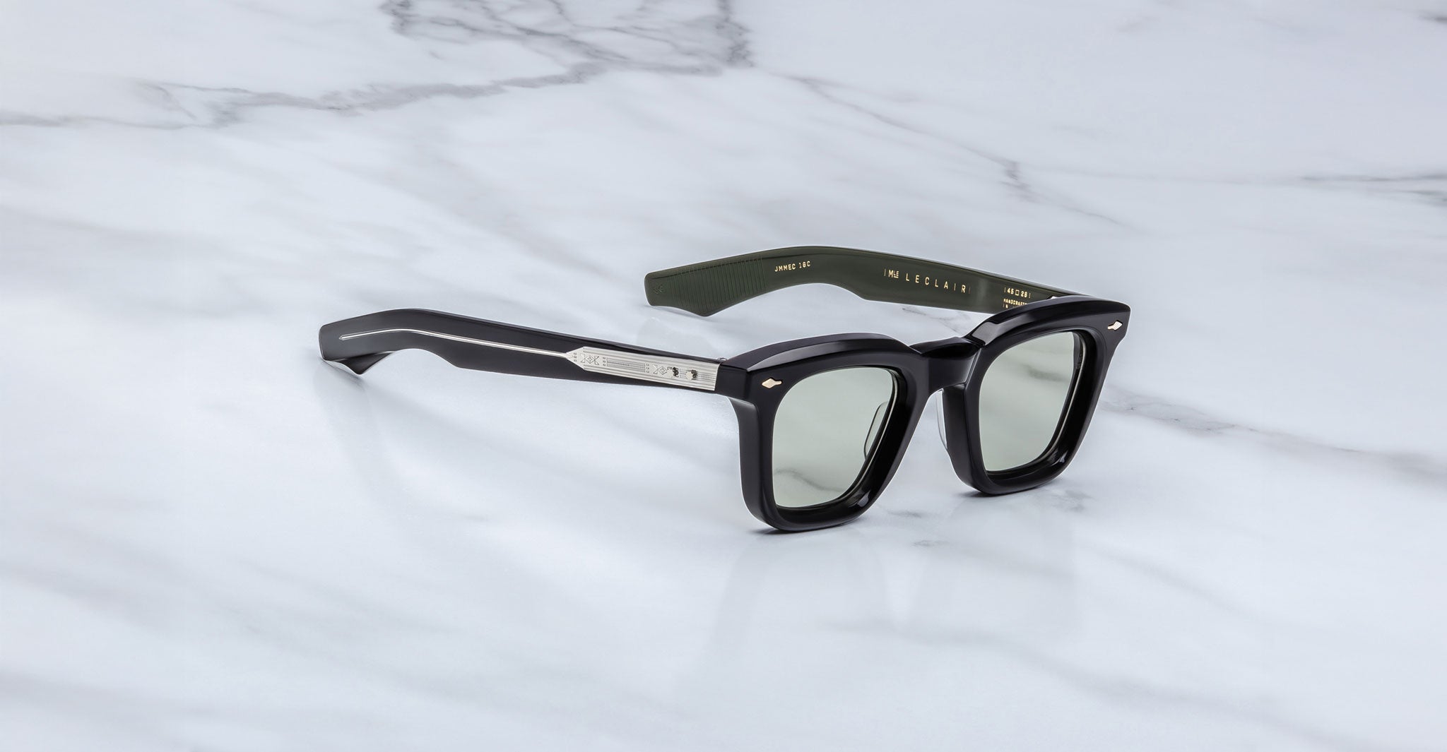 The Jacques Marie Mage Leclair sunglasses feature black rectangular frames with silver temple accents, tinted lenses, and gold text on the inner arms, displayed on a white marble surface.