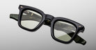 The Jacques Marie Mage Leclair eyeglasses feature thick-rimmed, square black frames with slightly tinted lenses, subtle ridged arms, and gold accents, displayed against a light gray background.