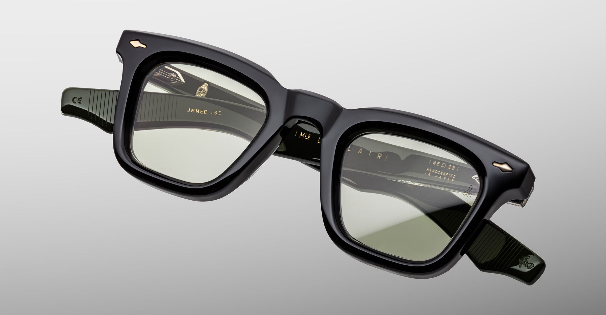 The Jacques Marie Mage Leclair eyeglasses feature thick-rimmed, square black frames with slightly tinted lenses, subtle ridged arms, and gold accents, displayed against a light gray background.
