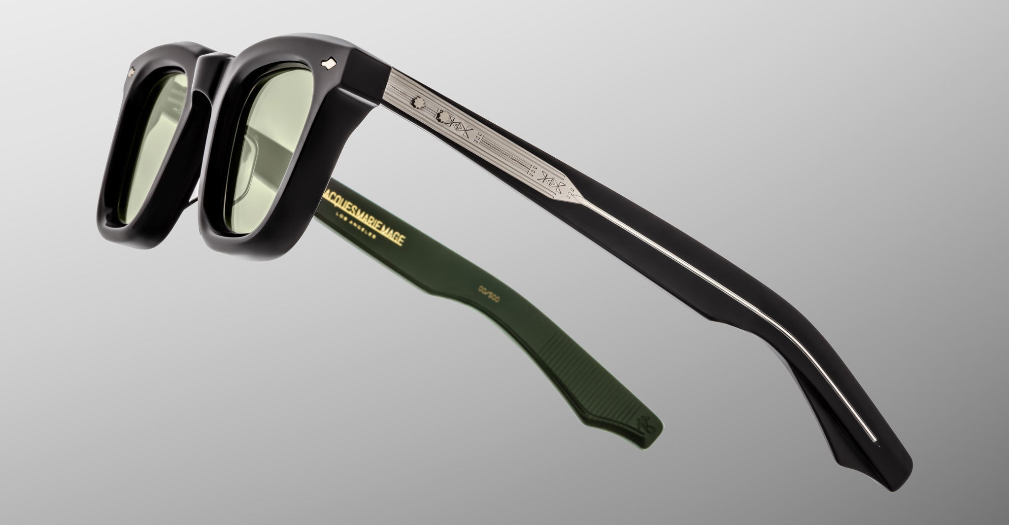Jacques Marie Mage Leclair sunglasses with black frames, green-tinted lenses, and green inner arms are shown angled on a grey background. The arm has a silver metallic accent and engraved branding.