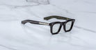 A pair of Jacques Marie Mage Leclair black rectangular eyeglasses with silver accents on the arms rests on a white marble surface. The inner left arm features dark green color and gold text.