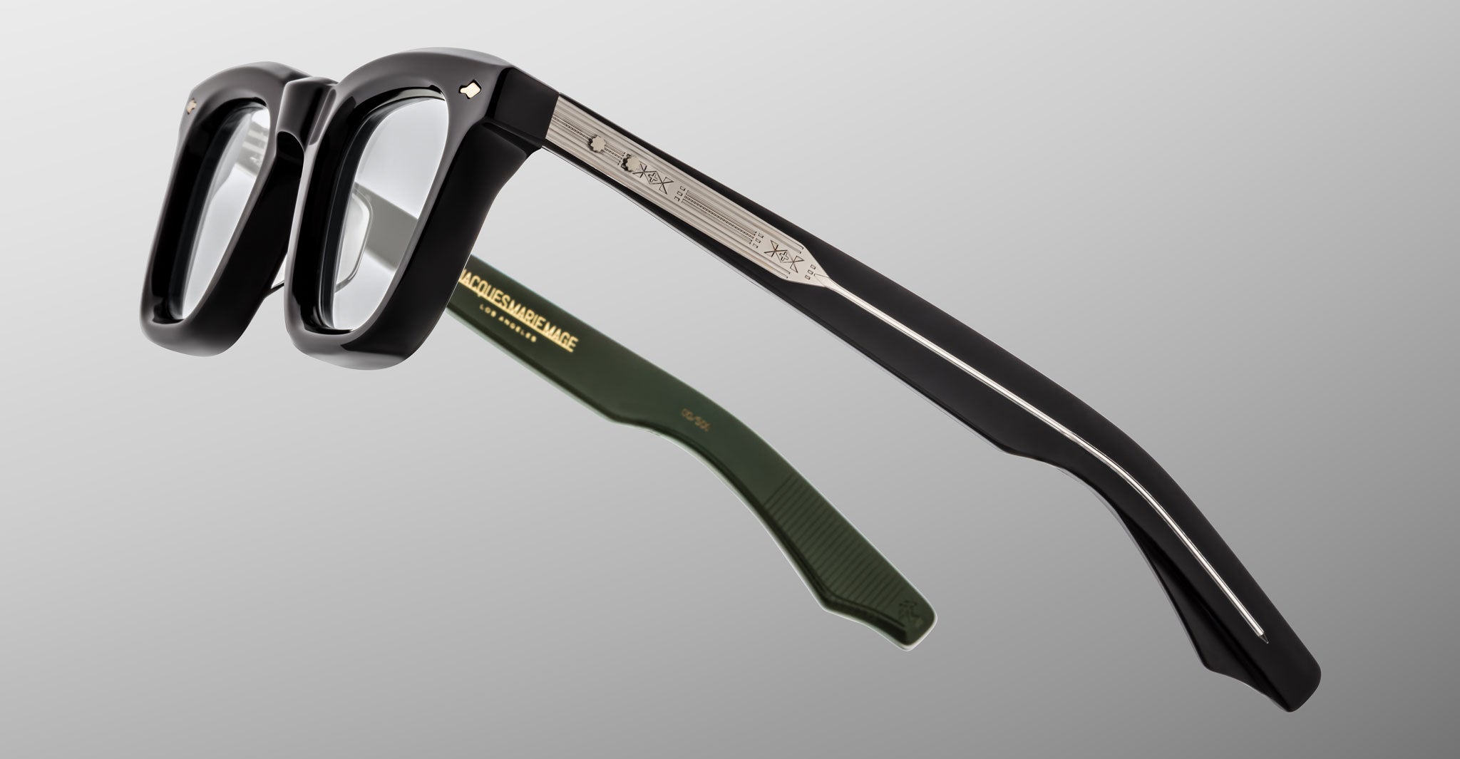A close-up of Jacques Marie Mage Leclair black eyeglasses with silver metal temples and green inner arms, featuring engraved details and a gold logo inside one arm, set against a gray background.