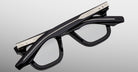 The Jacques Marie Mage Leclair eyeglasses, featuring thick black frames and silver engraved temples, are folded upside down on a light gray surface.