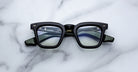 A pair of Jacques Marie Mage Leclair black rectangular eyeglasses with green arms and silver accents rests on a white marble surface, showcasing the brand name and model details inside the frame.