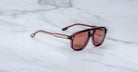 The Jacques Marie Mage Leonard sunglasses feature a burgundy frame with gold accents and tinted lenses, shown resting on a white marble surface.