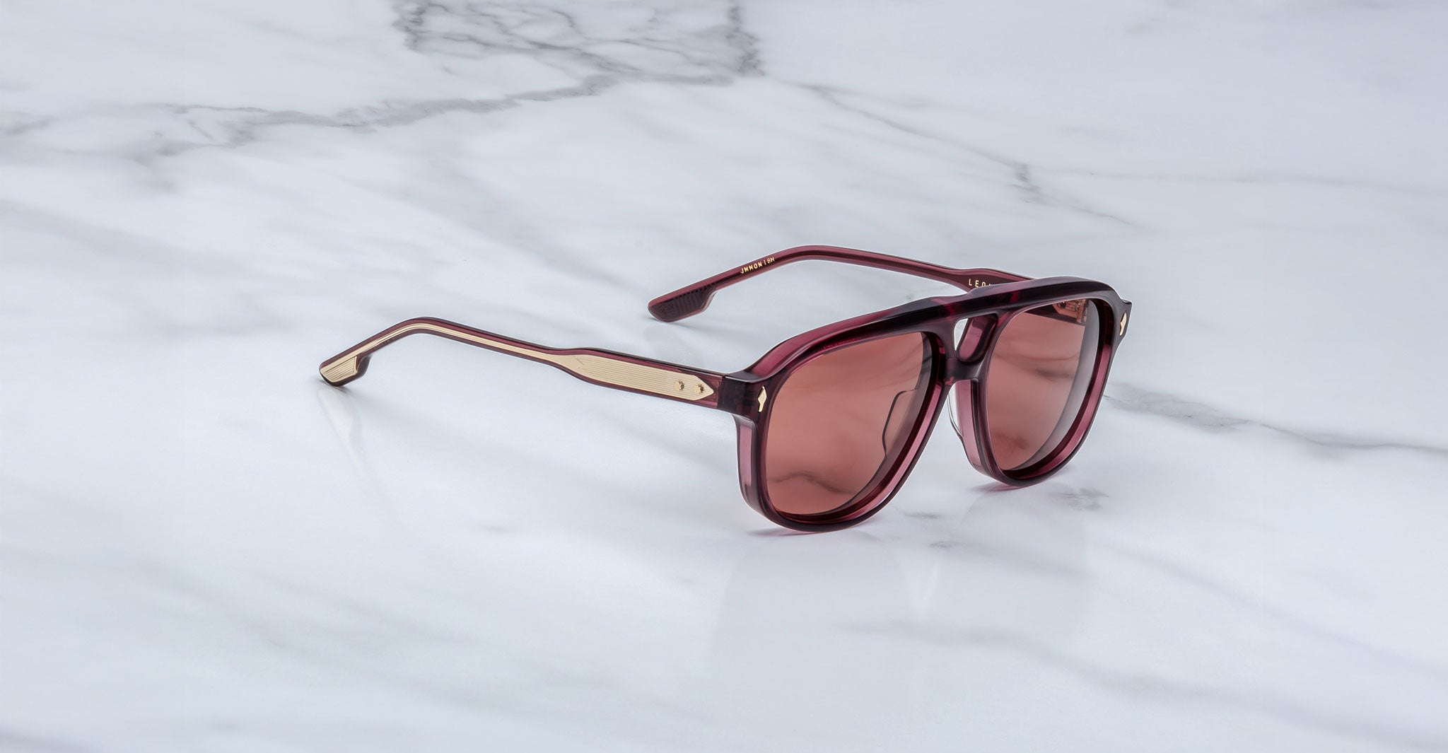 The Jacques Marie Mage Leonard sunglasses feature a burgundy frame with gold accents and tinted lenses, shown resting on a white marble surface.