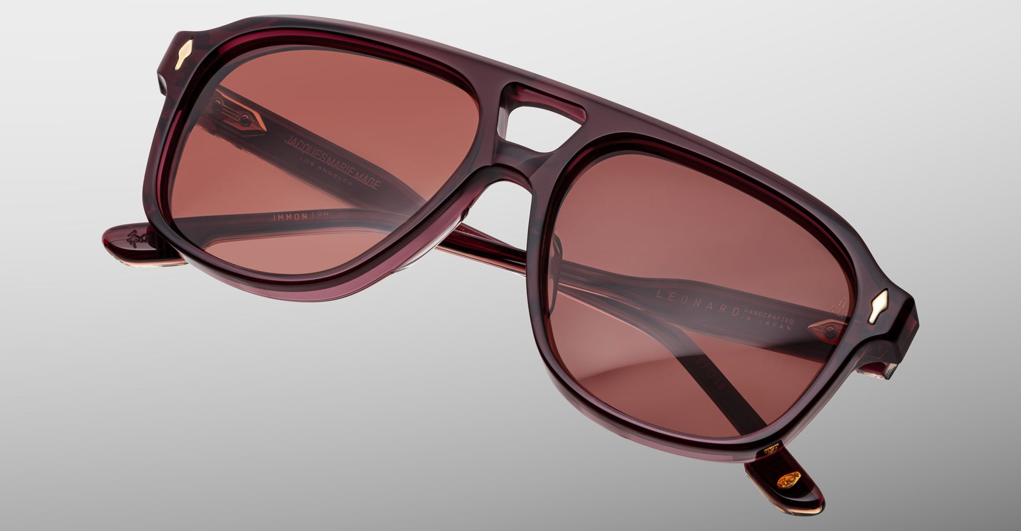 The Jacques Marie Mage Leonard sunglasses feature a dark red acetate frame with brown-tinted lenses, a keyhole bridge, and subtle metallic accents at the corners, displayed on a plain light gray background.