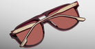 The Jacques Marie Mage Leonard sunglasses, featuring burgundy frames and rose-tinted lenses, are displayed upside down with gold arms accented in burgundy crossed over the lenses against a plain gradient background.