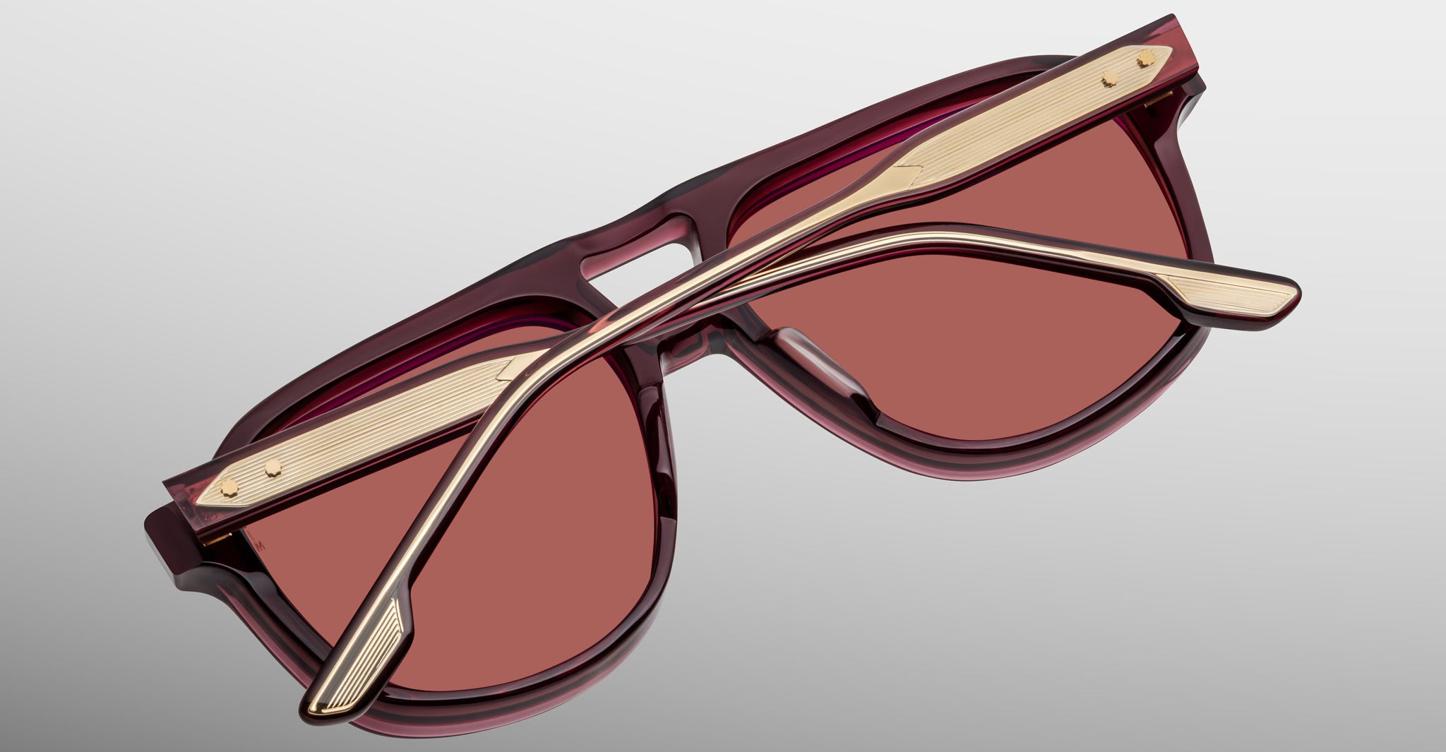 The Jacques Marie Mage Leonard sunglasses, featuring burgundy frames and rose-tinted lenses, are displayed upside down with gold arms accented in burgundy crossed over the lenses against a plain gradient background.