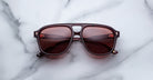 A pair of Jacques Marie Mage Leonard burgundy aviator-style sunglasses with red-tinted lenses rests on a white marble surface, showcasing branding and text on the arms.