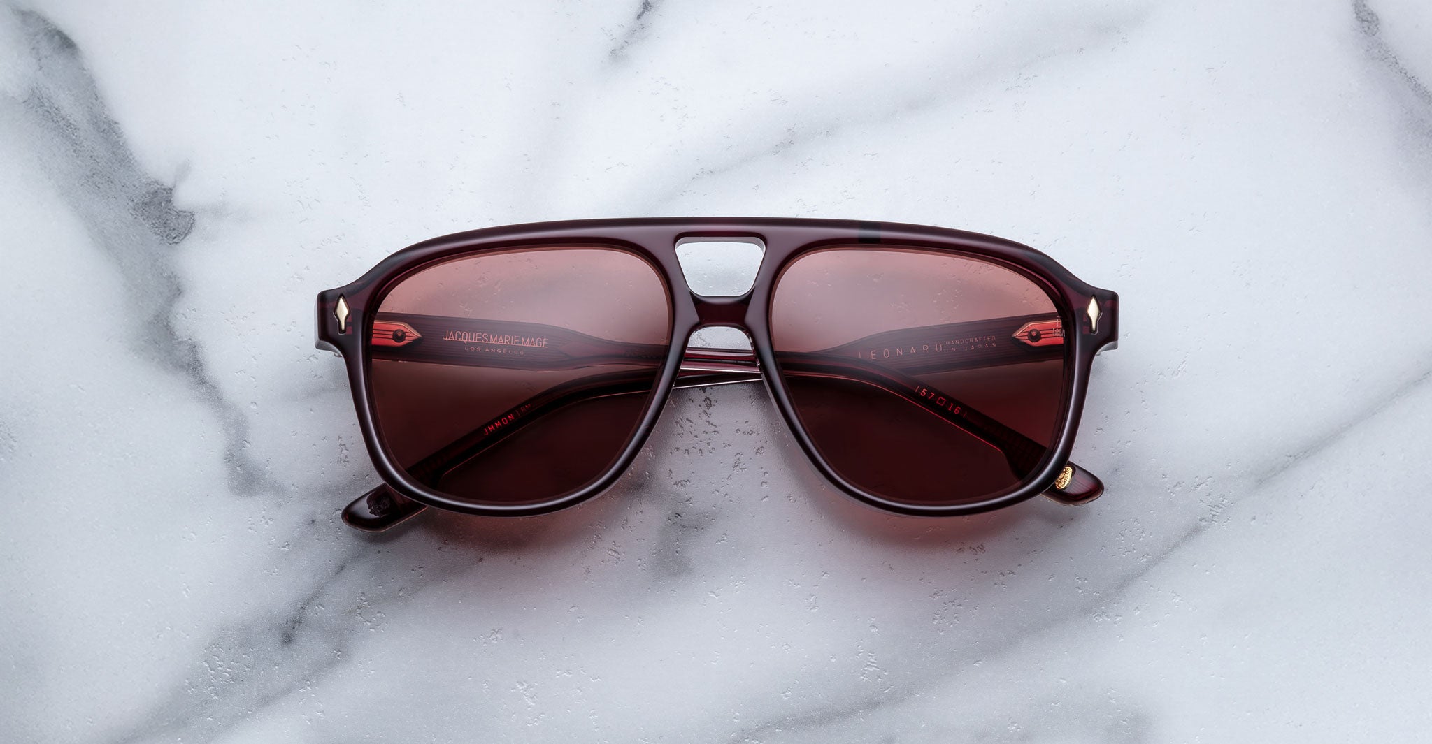 A pair of Jacques Marie Mage Leonard burgundy aviator-style sunglasses with red-tinted lenses rests on a white marble surface, showcasing branding and text on the arms.