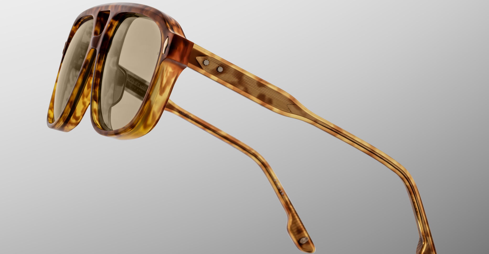 A close-up of Jacques Marie Mage Leonard sunglasses featuring a tortoiseshell pattern and brown-tinted lenses, photographed against a plain, light gray background.