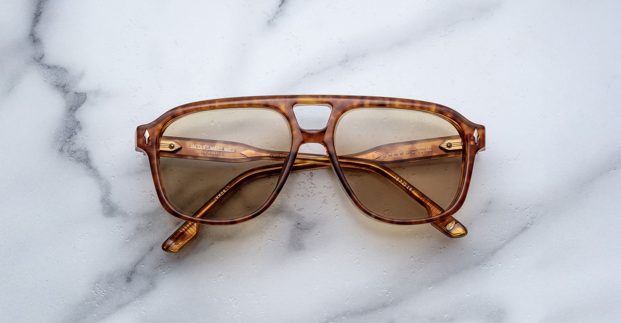 A pair of Jacques Marie Mage Leonard brown tortoiseshell sunglasses with light brown lenses is placed on a white marble surface.