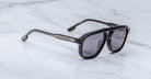 Jacques Marie Mage Leonard black sunglasses with thick frames and dark lenses rest on a light gray marble surface. The temples have metallic accents with two small rivets near the hinges.