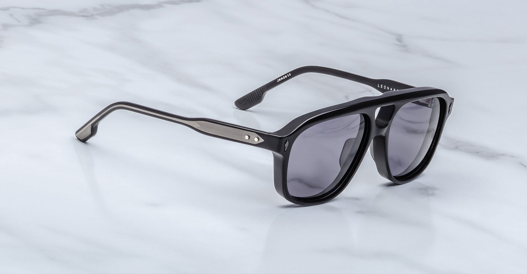 Jacques Marie Mage Leonard black sunglasses with thick frames and dark lenses rest on a light gray marble surface. The temples have metallic accents with two small rivets near the hinges.