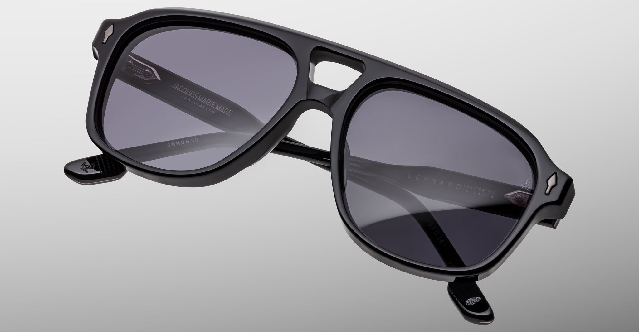A pair of black Jacques Marie Mage Leonard sunglasses with thick frames and dark lenses rests on a gray surface. The brand and model name are visible inside the temple arms.