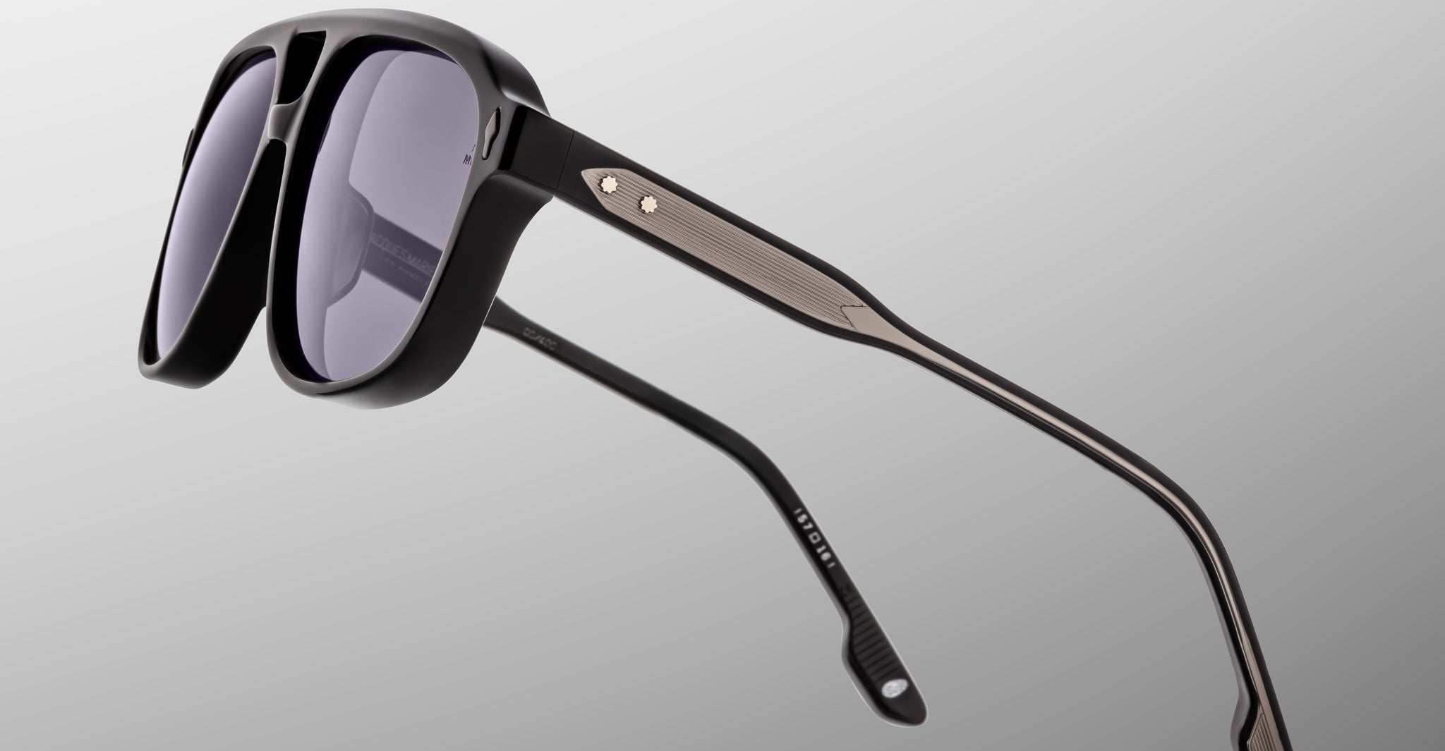 The Jacques Marie Mage Leonard sunglasses feature black frames, dark tinted lenses, and thin, textured metal arms with a small round detail by the right hinge, displayed against a light gray background.
