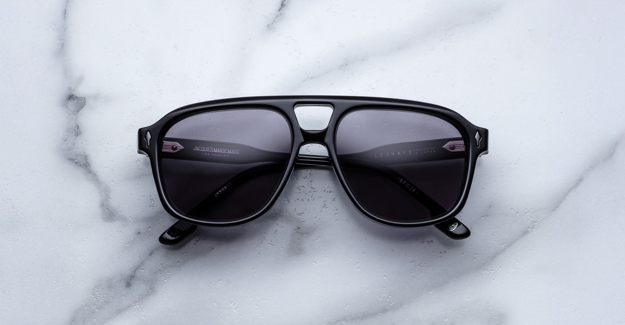 A pair of Jacques Marie Mage Leonard sunglasses with black frames and dark lenses rests on a white marble surface, shown from above with both lenses and arms clearly visible.