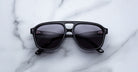 A pair of Jacques Marie Mage Leonard sunglasses with black frames and dark lenses rests on a white marble surface, shown from above with both lenses and arms clearly visible.