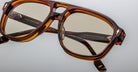 Close-up of Jacques Marie Mage Leonard eyeglasses featuring a stylish brown tortoiseshell design, square lenses, gold accents, and inner-arm text, set against a light gray background.