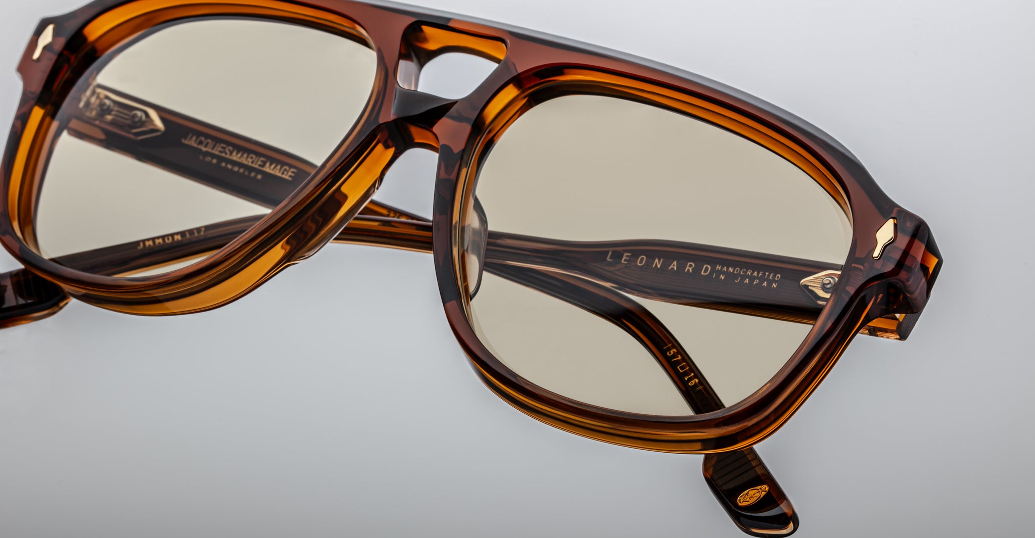 Close-up of Jacques Marie Mage Leonard eyeglasses featuring a stylish brown tortoiseshell design, square lenses, gold accents, and inner-arm text, set against a light gray background.