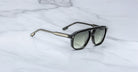 The Jacques Marie Mage Leonard sunglasses, featuring tinted lenses and a slightly angular black frame with silver arm accents, are displayed on a white marble surface.