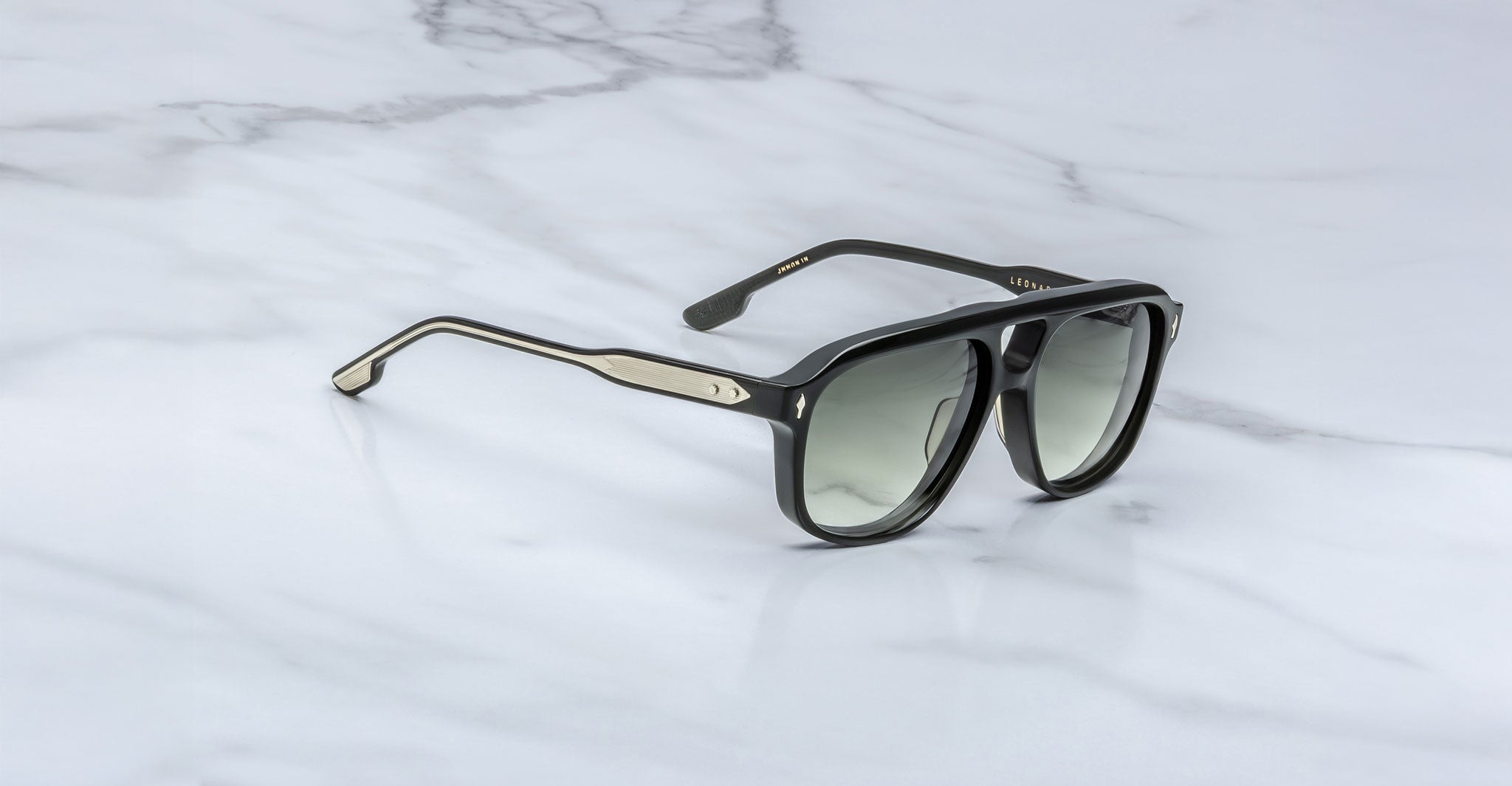 The Jacques Marie Mage Leonard sunglasses, featuring tinted lenses and a slightly angular black frame with silver arm accents, are displayed on a white marble surface.