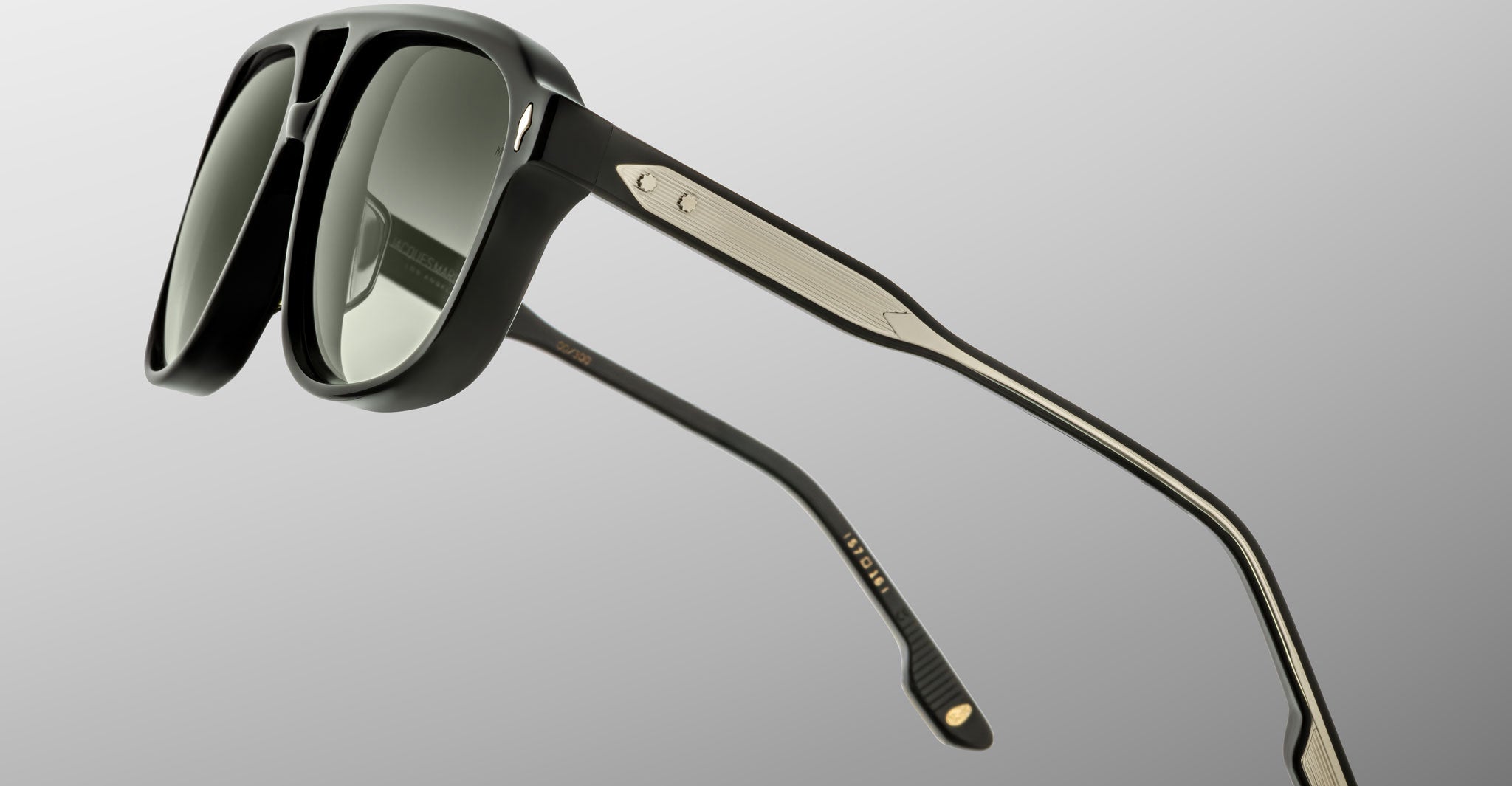 The Jacques Marie Mage Leonard sunglasses feature thick black frames, green-tinted lenses, metallic detailing on the arms, and subtle gold accents near the hinges, set against a plain gradient background.